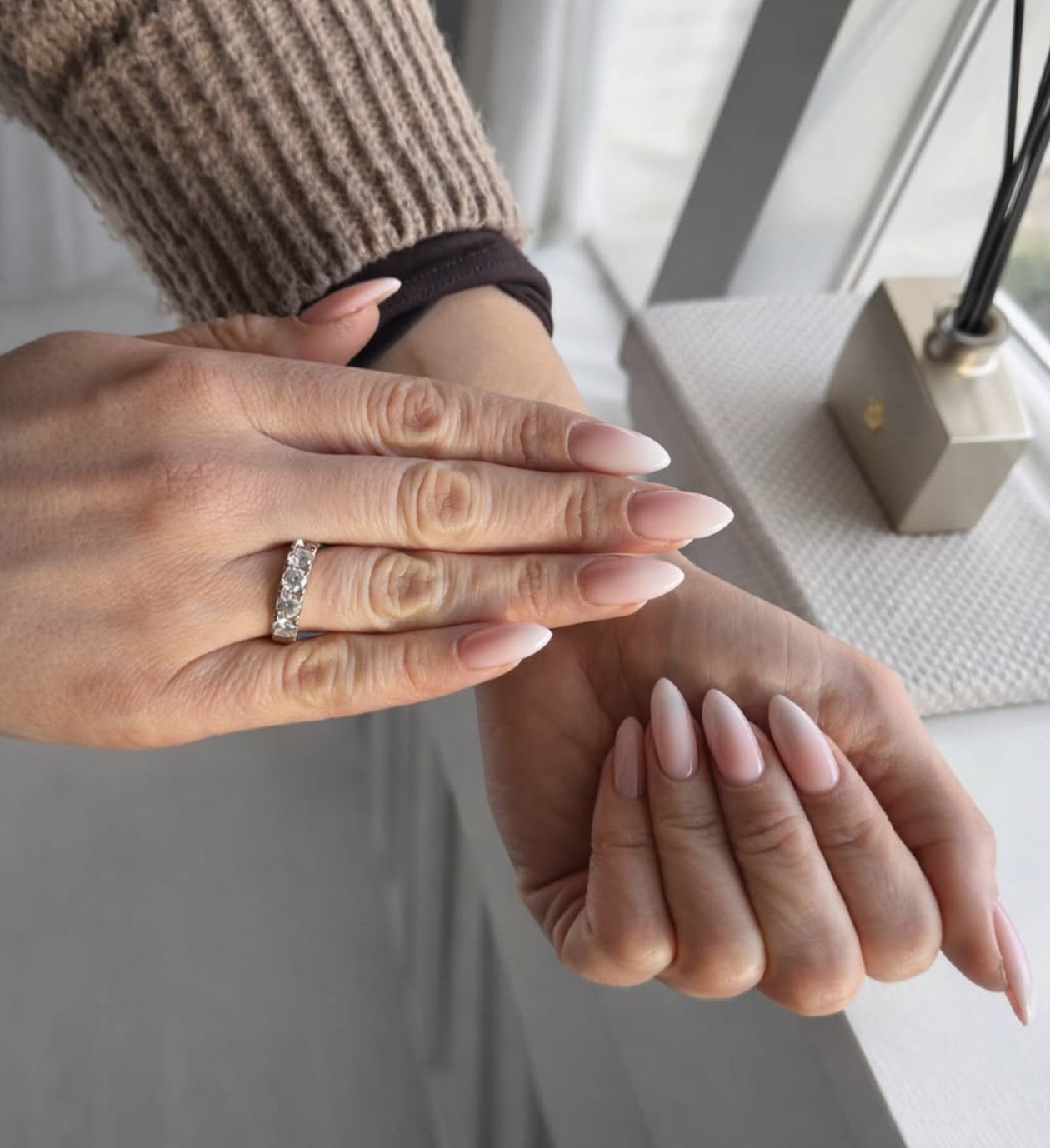 Twee handen met nude amandelvormige BIAB-nagels bij het raam